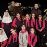 دانش‌آموزان دختر در یونیفرم مشکی و صورتی در حال اجرای صحنه‌ای، با معلمان و تزئینات درختی در پس‌زمینه