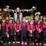گروهی از دانش‌آموزان با لباس مشکی و صورتی در کنار معلم، عروسک‌های تئاتری بالای سر دارند، در حال اجرای نمایش تئاتری روی صحنه