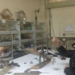 معلم و دو دانش‌آموز در حال انجام فعالیت و آزمایش علمی در آزمایشگاه مدرسه با قفسه‌های تجهیزات و ابزار آزمایشگاهی