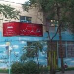 نمای بیرونی ساختمان مدرسه با درب ورودی و تابلوی قرمز و دیوار آبی‌رنگ