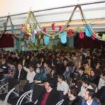 تجمع دسته‌جمعی از دانش‌آموزان و افراد در یک سالن مزیّن شده با بالاهای کاغذی رنگین و تزئینات رنگارنگ، به نظر مراسم یا جشن مدرسه