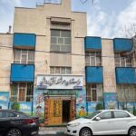 نمای جلویی ساختمان مدرسه با تابلوی نام دائمی فارسی بالای درب ورودی، درب طلایی زیبا، پنجره‌های آبی، و معماری معاصری