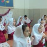 دانش‌آموزان دختر با لباس مدرسی و حجاب سفید در کلاس درس نشسته‌اند. تابلو رنگی و پنجره‌های روشن در پس‌زمینه دیده می‌شود.