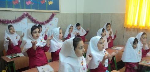 دانش‌آموزان دختر با لباس مدرسی و حجاب سفید در کلاس درس نشسته‌اند. تابلو رنگی و پنجره‌های روشن در پس‌زمینه دیده می‌شود.