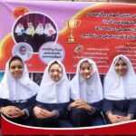چهار دانش‌آموز دختر با پوشش فرم مدرسه و حجاب سفید در حال شرکت در مسابقه‌ی فرهنگی مدرسه، جلوی بنر تبلیغاتی رویداد