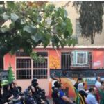 گردهمایی دانش‌آموزان و کارکنان مدرسه در ورودی ساختمان با دیوار قرمز رنگ، احتمالاً برای یک مراسم یا رویداد مدرسه‌ای
