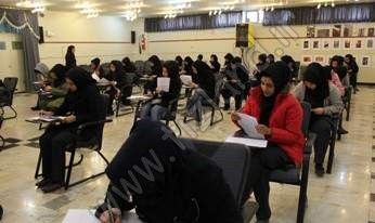 دانش‌آموزان در حال شرکت در یک جلسه آزمون گروهی در سالن، نشسته در میزهای مختلف