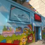 نمای بیرونی ساختمان مدرسه با درب ورودی آبی‌رنگ و تابلو نام‌نوشته طوره روی دیوار