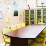 کلاس درس علوم با میز بیضی‌شکل سفید، صندلی‌های زرد، کابینت‌های سبز پر از تجهیزات آزمایشگاهی و پوسترهای آموزشی علمی بر روی دیوار