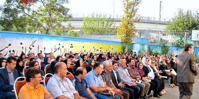 دانش‌آموزان و معلمان مدرسه در حال شرکت در یک مراسم یا اجلاس رسمی در فضای درون‌محوطه‌ای، با پس‌زمینه ساختمان‌های مدرسه و تابلوی رنگین.