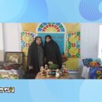 دو دانش‌آموز در کنار میز نمایشی با تابلوهای هنری رنگی و تزیینات در پس‌زمینه