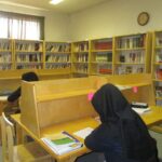 کتابخانه مدرسه با قفسه‌های کتاب زرد رنگ، میزهای مطالعه و دانش‌آموزان در حال مطالعه