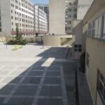 حیاط باز مدرسه با زمین سیمانی، ساختمان‌های مسکونی دوری، و درخت سبز