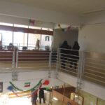 نمای کلی از راهروی مدرسه با نرده‌های فلزی، نورگیرهای بزرگ، و دکوراسیون‌های رنگی