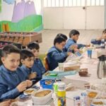دانش‌آموزان در حال خوردن ناهار در سالن غذاخوری مدرسه، نشسته دور میز با ظروف غذا و مشروبات