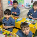 چهار دانش‌آموز جوان در حین انجام فعالیت‌های یادگیری با پازل‌های آموزشی و بازی‌های رنگی روی میز‌های زرد در کلاس درس