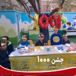 دانش‌آموزان درحال برپایی ایستگاه فعالیت در یک رویداد مدرسه، با میز تزیین شده، بادکنک‌های قرمز شماره 100 و کادوهای رنگی