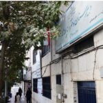 نمای خارجی ساختمان مدرسه با درب ورودی آبی و تابلوی نام‌گذاری رسمی به رنگ آبی و سفید