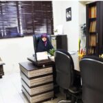 اتاق کامپیوتر مدرسه با میز‌های کار، صندلی‌های سیاه اداری، کامپیوتر، و قفسه‌های کتابی دیواری، روشن‌شده از نور طبیعی پنجره