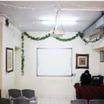 نمای درونی کلاس درس با تخته سفید، کتابخانه‌های چوبی، تزیینات گیاهی آویزان، صندلی‌ها و تصاویر دیواری