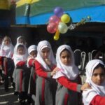 دانش‌آموزان دختر با لباس یکدست قرمز و سفید در حال شرکت در جشن یا مراسم گروهی در حیاط مدرسه با تزیین بادکنک‌های رنگی