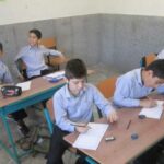 دانش‌آموزان پسر در حال درس خواندن و نوشتار در کلاس درس با میز‌های آبی و صندلی‌های نارنجی