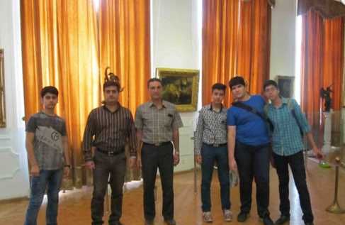 گروهی از معلمان و مسئولان مدرسه در حال حضور در یک نمایشگاه یا رویداد رسمی داخل فضای گالری با دیوارهای سفید، پردهای طلایی و آثار هنری