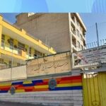 نمای بیرونی درب ورودی اصلی مدرسه با ساختمان آجری چند طبقه‌ای، درب رنگی و تابلوی فارسی