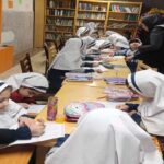دانش‌آموزان در حال مطالعه کتاب در کتابخانه مدرسه با قفسه‌های پر از کتاب‌های مختلف