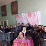 گروهی از دانش‌آموزان و اساتید در حال شرکت در مراسم جشن مدرسه‌ای با تزیینات گل کاغذی صورتی و سفید