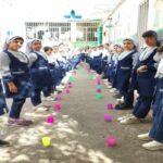گروه بزرگی از دانش‌آموزان دختر در لباس فرم تیره در حیاط مدرسه، شرکت‌کننده در یک فعالیت بازی‌گونه با اشیاء رنگی روی زمین