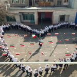 دانش‌آموزان با لباس سفید در حیاط مدرسه حول پرچم‌های ایران در یک مراسم رسمی ایستاده‌اند