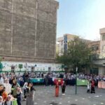 دانش‌آموزان در حیاط مدرسه حین جمع‌آوری یا برنامه‌ی فرهنگی، با دیوارهای نقاشی‌شده و تجهیزات بازی