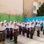 دانش‌آموزان دختر مدرسه در حیاط با لباس یونیفرم و روسری سفید در حال صف‌بندی منظم برای مراسم صبحگاهی
