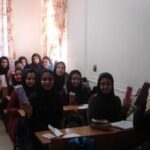 گروهی از دانش‌آموزان دختر نشسته و ایستاده در یک فصل درسی با پنجره‌های آفتاب‌گیر ستائر قرمز و لوح سفید