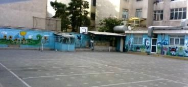 صحن و حیاط مدرسه با پوشش بتنی و ساختمان‌های آموزشی درحال‌حاضر در پس‌زمینه