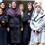گروهی از معلمان و دانش‌آموزان دختر ایستاده جلوی ورودی ساختمان مدرسه، با گل‌های رنگی در جلویشان، در یک عکس یادگاری گروهی
