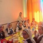 گروهی از دانش‌آموز دختر با روسری، نشسته در کنار میزی در حال شرکت در یک مراسم یا رویداد اجتماعی مدرسه‌ای با میان‌وعده‌ها