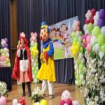 دو دختربچه با لباس فانتزی در مراسمی جشنواره با تزیینات بالون رنگارنگ، بنرهای تزیینی، و گل‌های مصنوعی