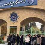 درب ورودی اصلی ساختمان آجری مدرسه با تابلوی آبی و نشان دائمی، دانش‌آموزان در جلوی درب‌های سبز