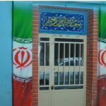 درب ورودی اصلی ساختمان مدرسه با تابلوی نام‌گذاری آبی و نمادهای ایرانی