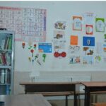 کلاس درسی برای آموزش انگلیسی با میز‌های یادگیری، کتابخانه کناری، و دیوار تزیین‌شده با تابلوی حروف و اعداد انگلیسی و طرح‌های رنگی آموزشی