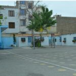 حیاط مدرسه با فرش خطی‌دار، درخت‌های سبز، و نمای ساختمان‌های آموزشی از پس‌زمینه