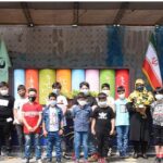 گروهی از دانش‌آموزان در حال شرکت در فعالیت آموزشی محیط‌زیست و پروژه نمایشگاهی با پس‌زمینه پنل‌های رنگین و پرچم ایران