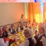 دختران دانش‌آموز با روسری سفید و لباس‌های رسمی دور میز زرد در سالن ناهارخوری، در حال خوردن غذا