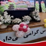 صحنه تزیین‌شده برای مراسم مدرسه با گل‌های سفید، بادکنک‌های رنگی و پرده‌های سیاه، و تابلویی با متن فارسی