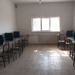 اتاق سایت کامپیوتر مجهز به رایانه‌های دسکتاپ و صندلی‌های چوبی، نور طبیعی از پنجره‌های بزرگ