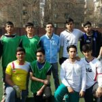 تصویر تیمی از ورزشکاران جوان در لباس تمرینی مختلف رنگ ایستاده در کنار میدان ورزشی محوطه‌ی مدرسه