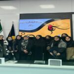 گروهی از دانش‌آموزان با لباس مشکی و اعضای کادر آموزشی در حال شرکت در مراسمی رسمی به پایِ پرچم ملی ایران و یک بنر تزئینی