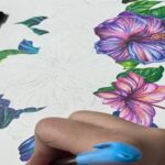 دانش‌آموزی درحال نقاشی گل‌های بنفش و صورتی با رنگ آبی روی کاغذ سفید، تصویری از فعالیت هنری درون کلاس درس